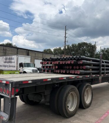 SGK Global Shipping Services truck hauling a flatbed of steel pipes past an orange shipping container in Houston